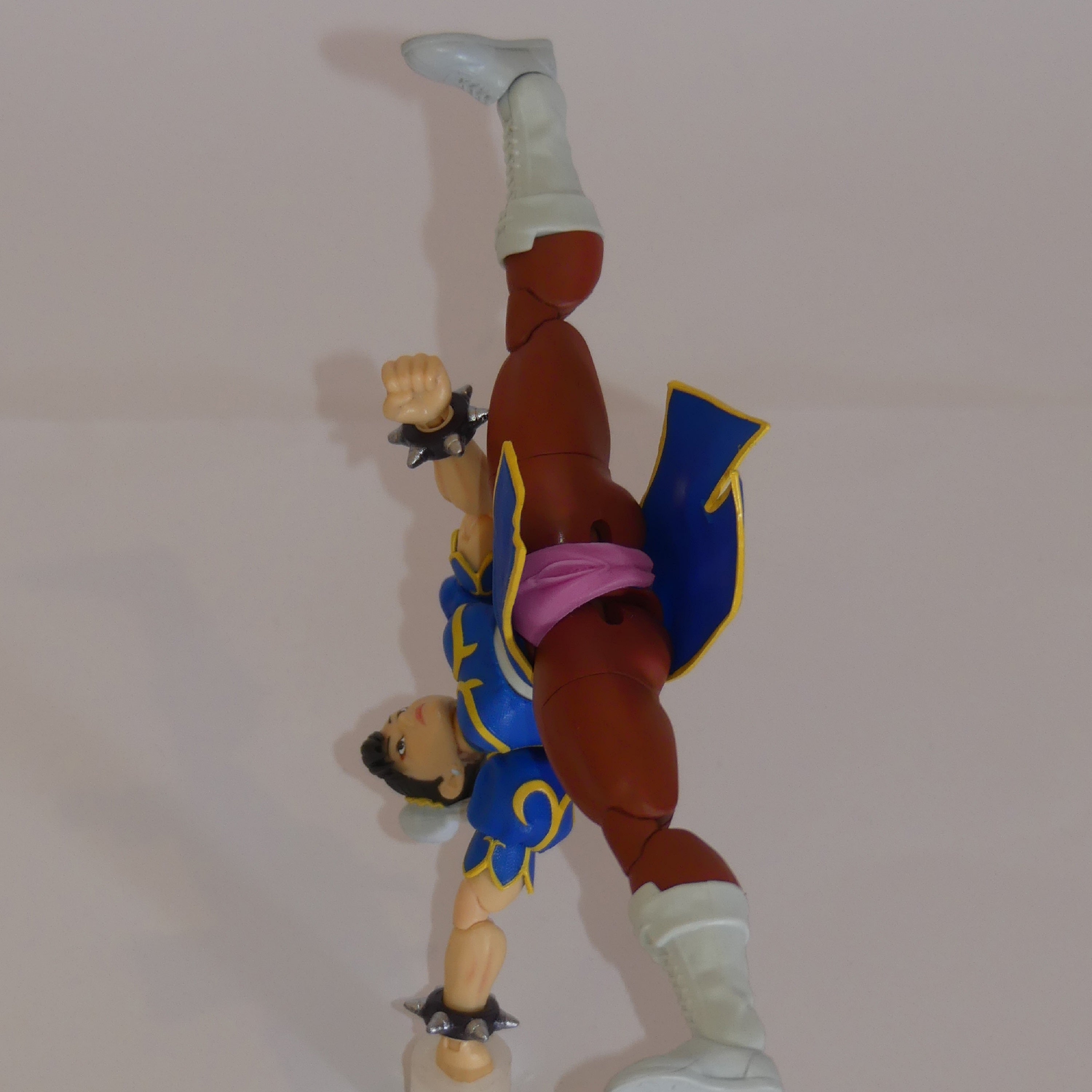 A Street Fighter action figure of Chun-Li posed dynamically in a handstand, wearing her signature blue and gold outfit with a pink silicone vulva vagina, white boots, and brown skin.