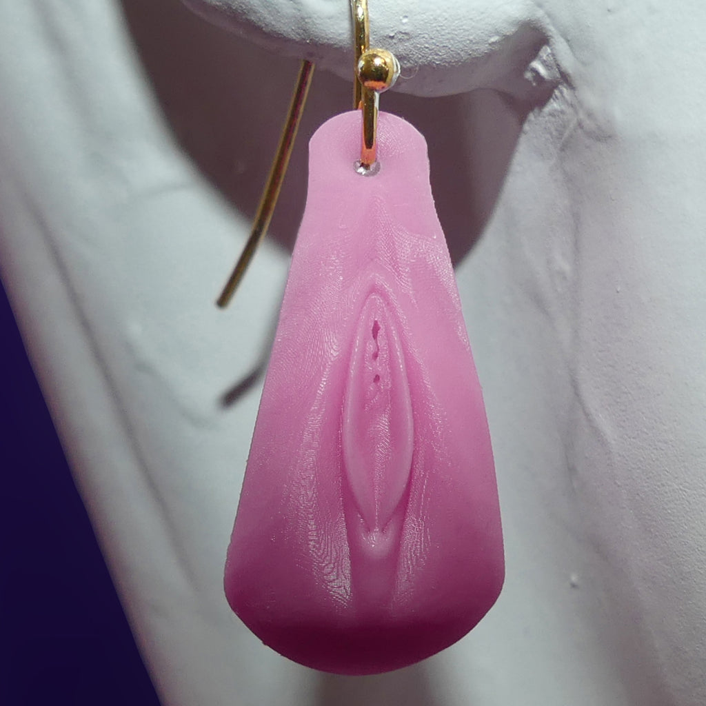 A pink silicone novelty dangle earring shaped like a stylized vulva, suspended from a gold-tone fishhook ear wire, displayed on a white mannequin bust.