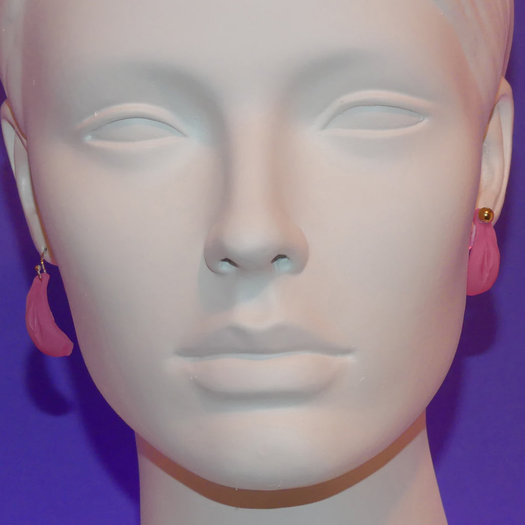 Pink crescent-shaped dangle earrings displayed on a white mannequin head against a purple background.