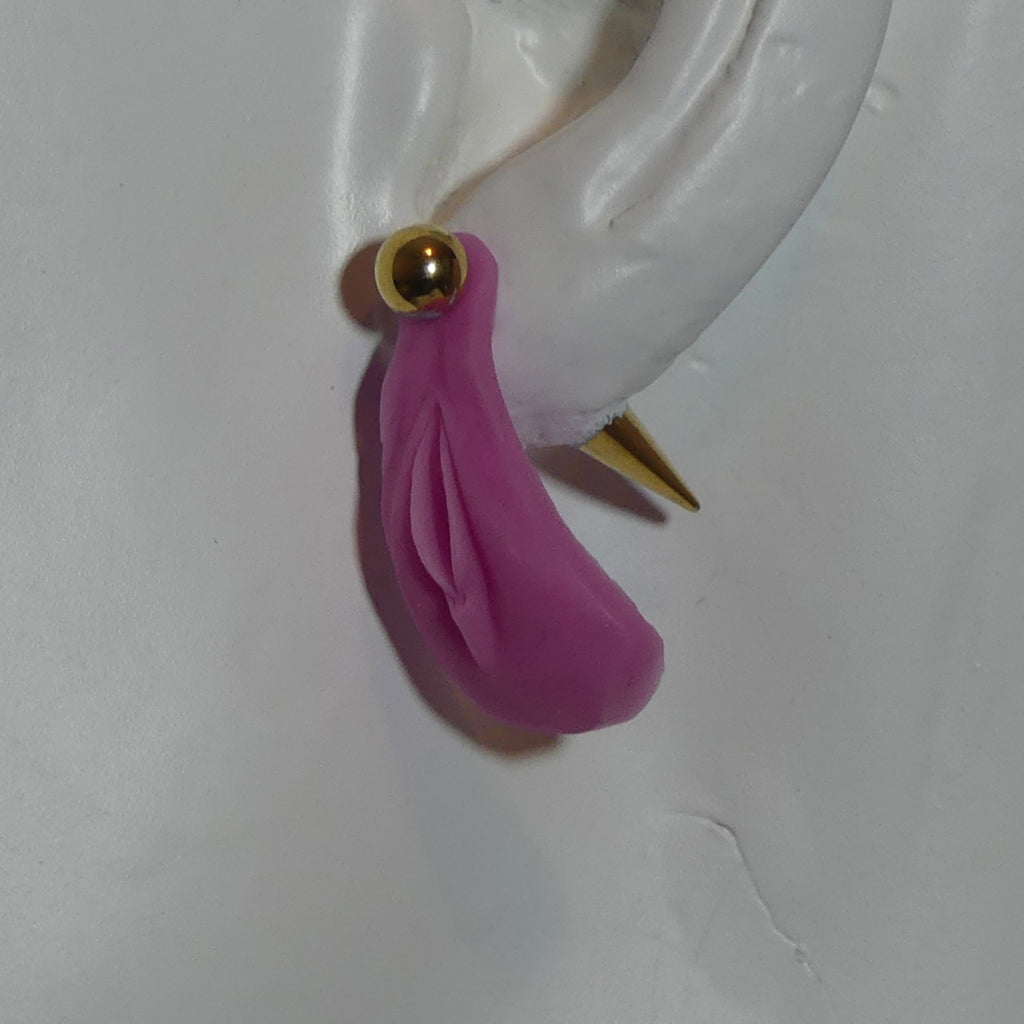 A pink vulva dangle earring with a gold ball stud and gold spike back, shown on a white ear form against a white background.