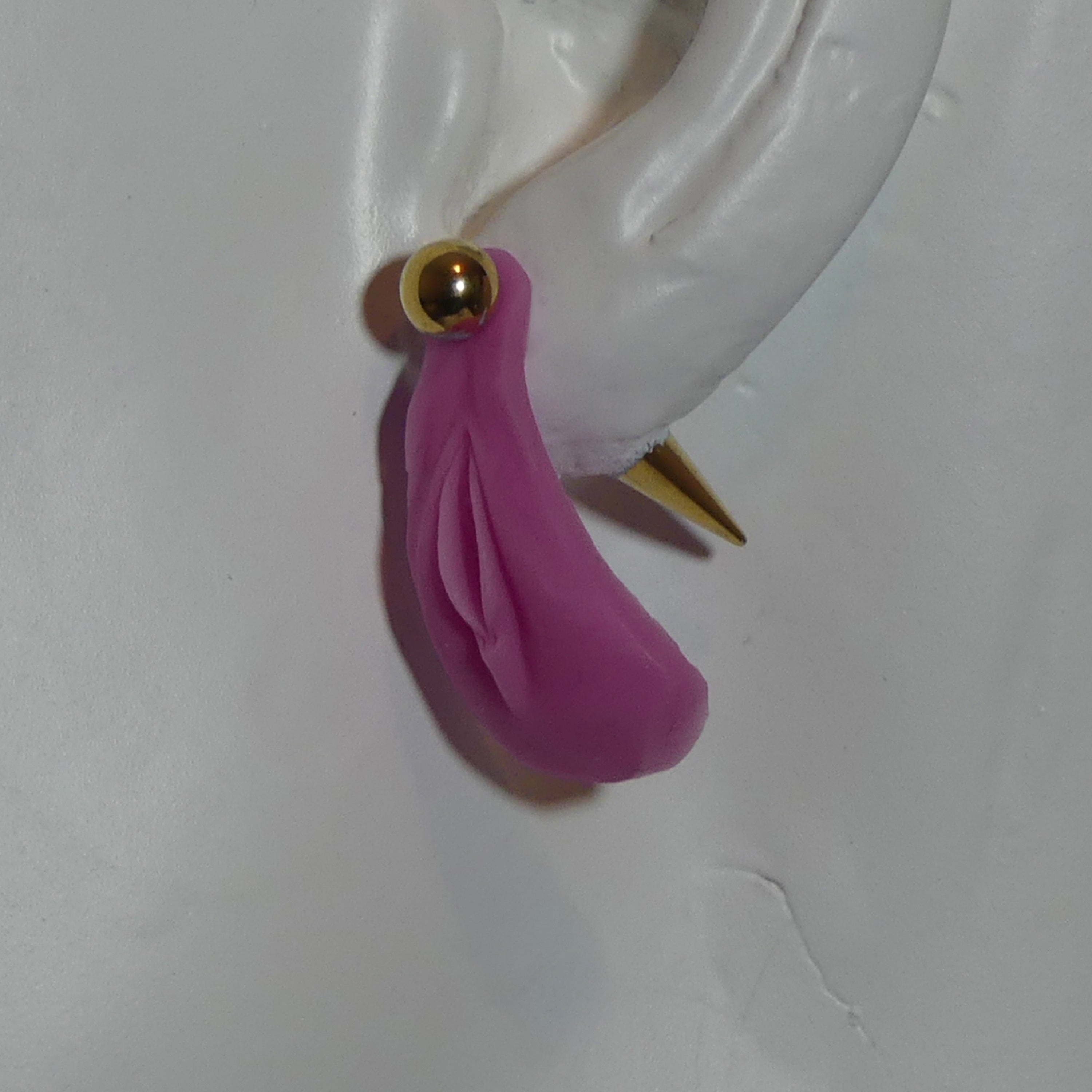 A pink vulva dangle earring with a gold ball stud and gold spike back, shown on a white ear form against a white background.