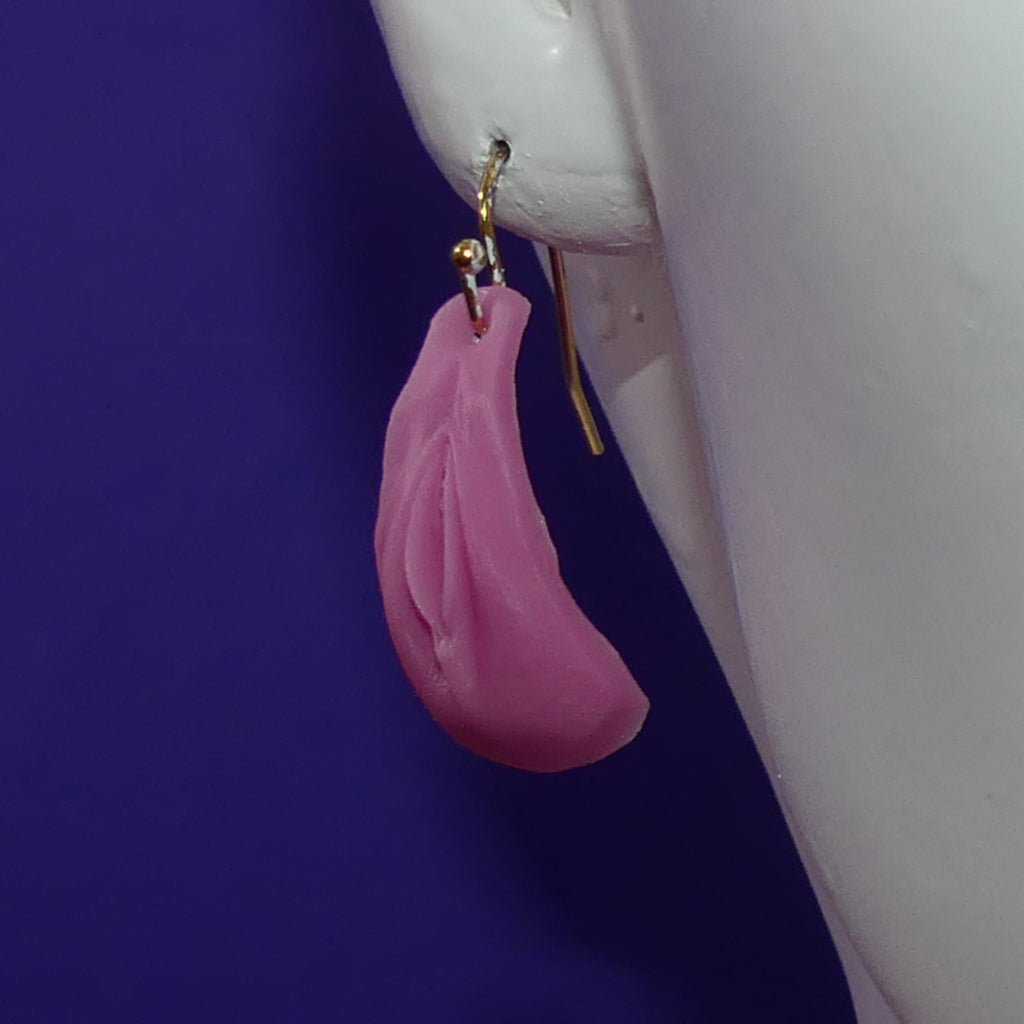 A single pink vulvar vagina drop earring with a gold fishhook clasp, shown hanging from a white mannequin ear against a solid dark purple background.
