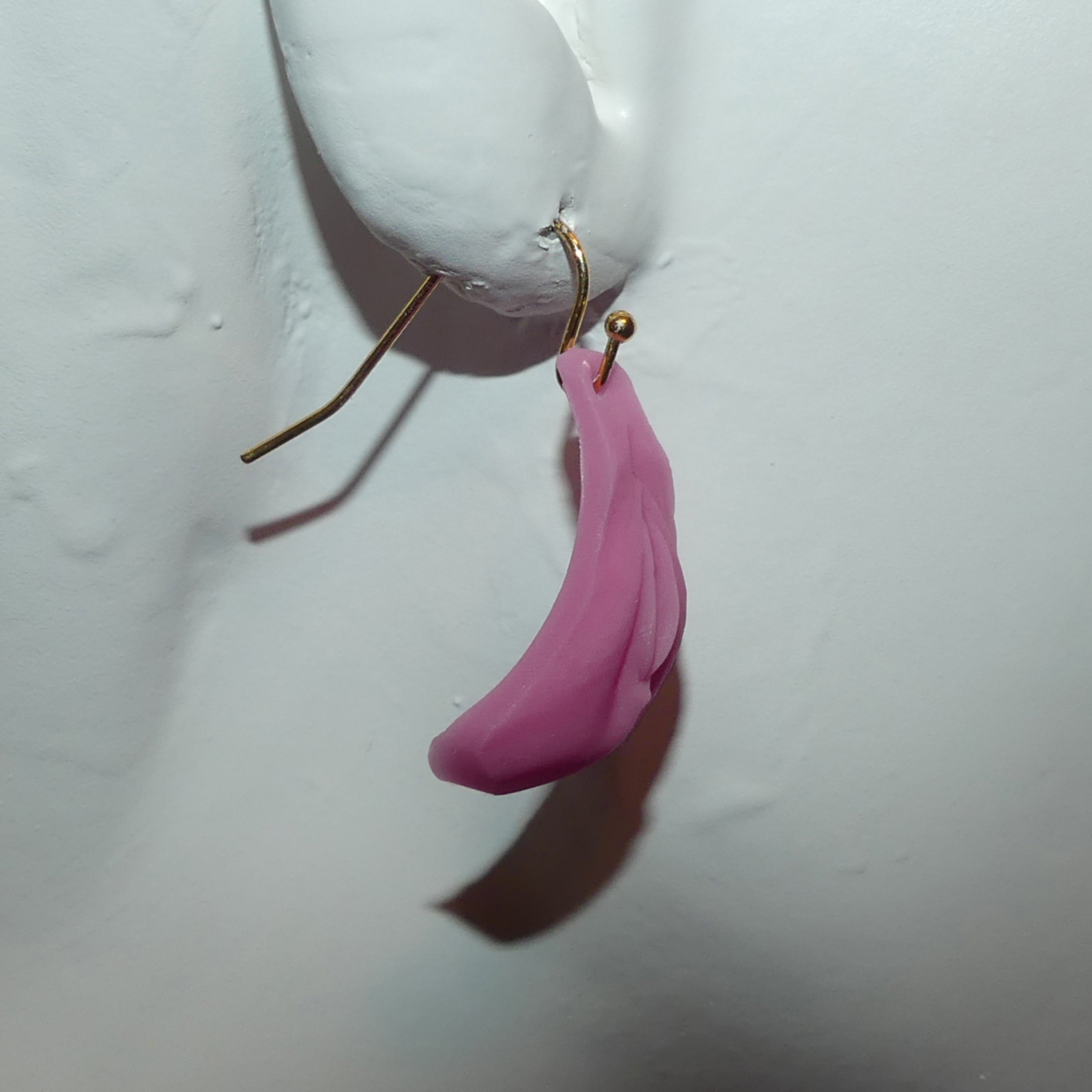 A single pink vulva-shaped dangle earring with a gold french hook, shown hanging against a white textured mannequin.