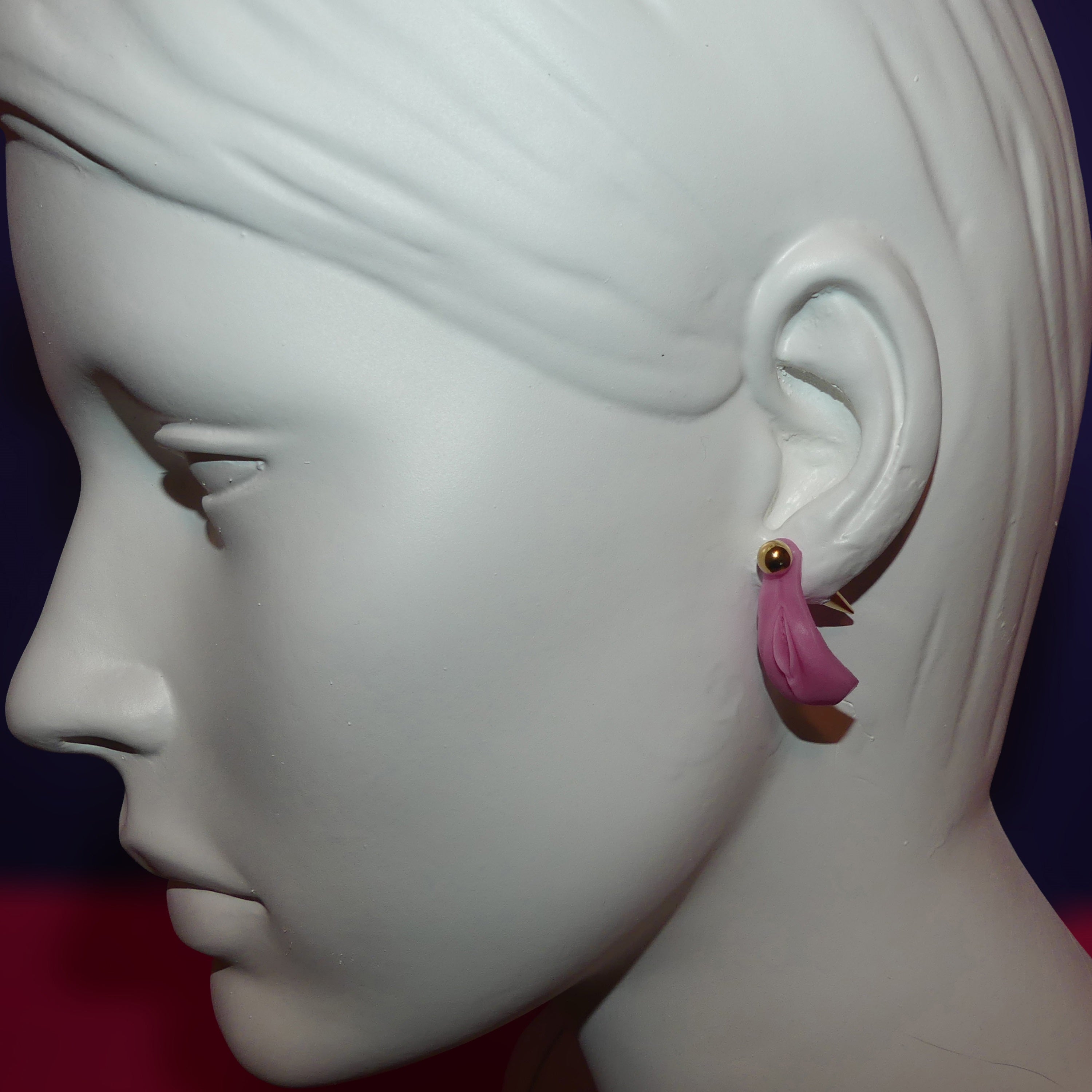 A pink and gold vagina-shaped dangle earring displayed on the ear of a white mannequin head in profile against a dark blue and red background.
