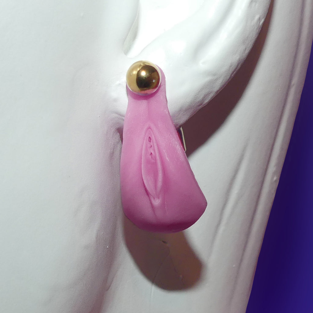 A pink silicone novelty earring shaped like a stylized vulva with a gold-tone ball stud, shown attached to a white mannequin ear.