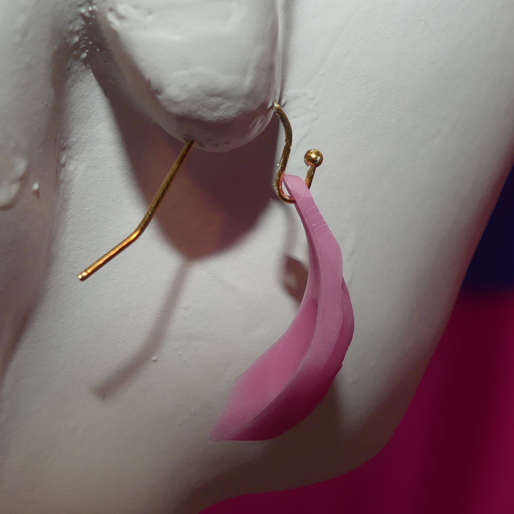 A close-up of a pink vagina-shaped dangle earring with a gold hook, displayed on a white mannequin bust against a pink background.