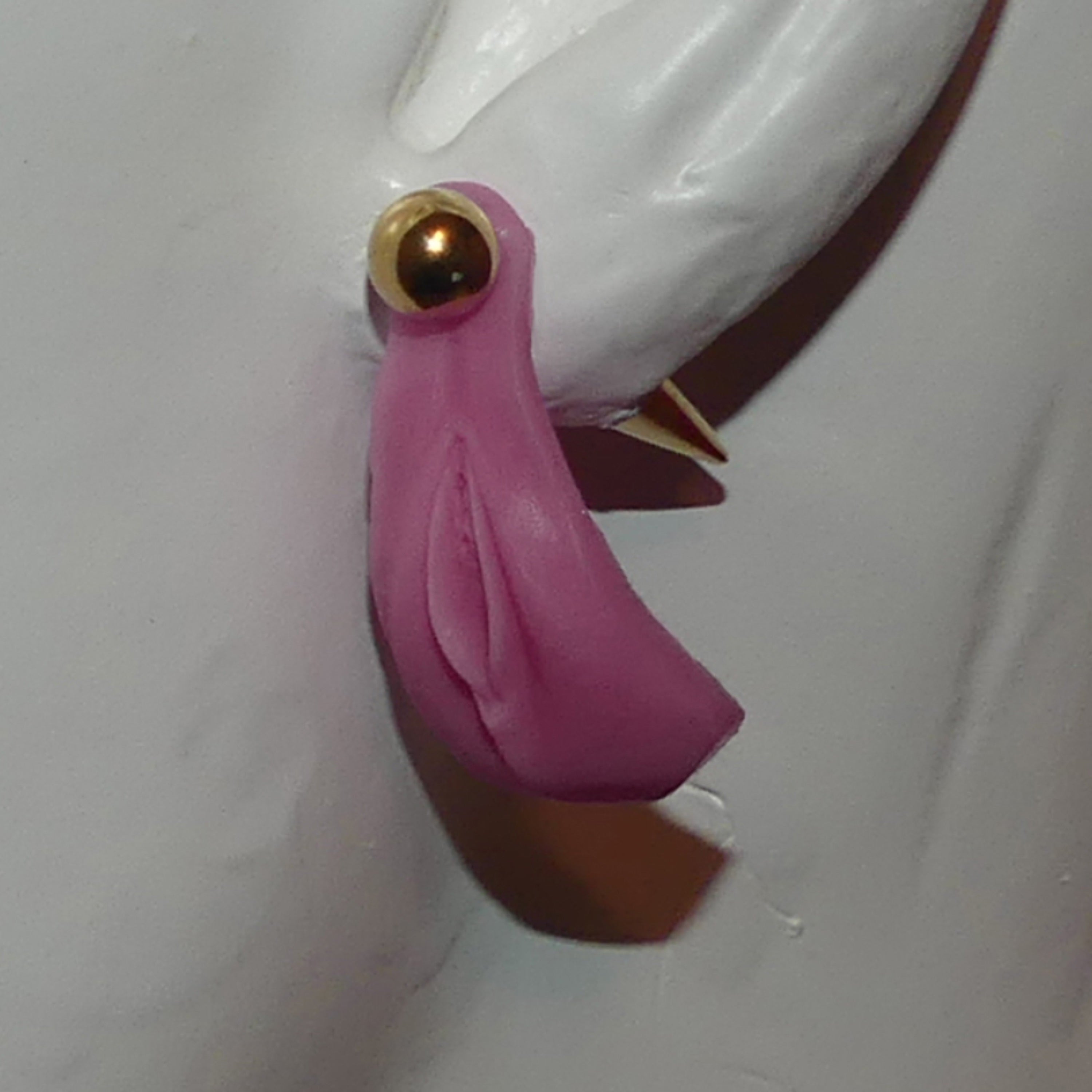 A close-up of a pink silicone vagina earring with a gold-toned circular stud and a small pointed gold accent, attached to a white sculptural form against a light gray background.