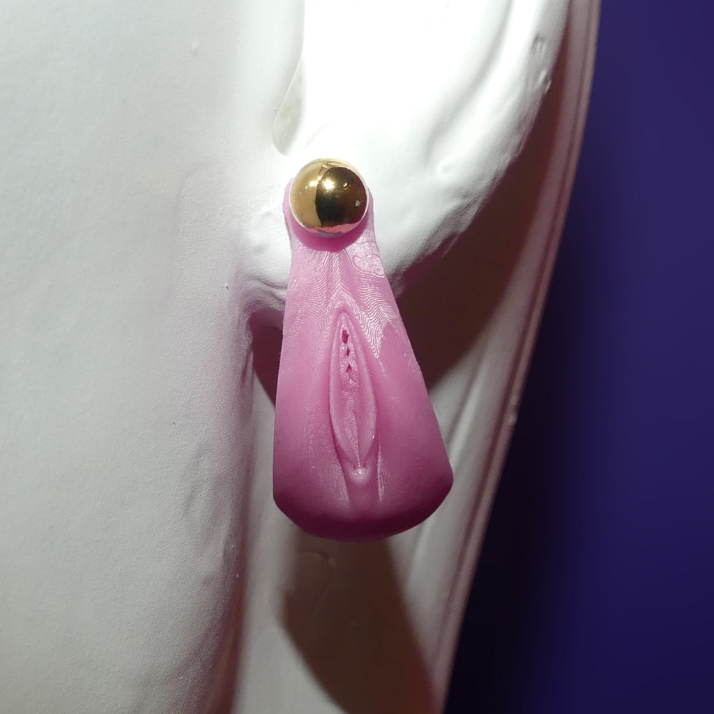 A pink silicone novelty stud earring shaped like a stylized vulva, featuring a gold-tone ball stud at the top, displayed on a white mannequin against a dark purple background.