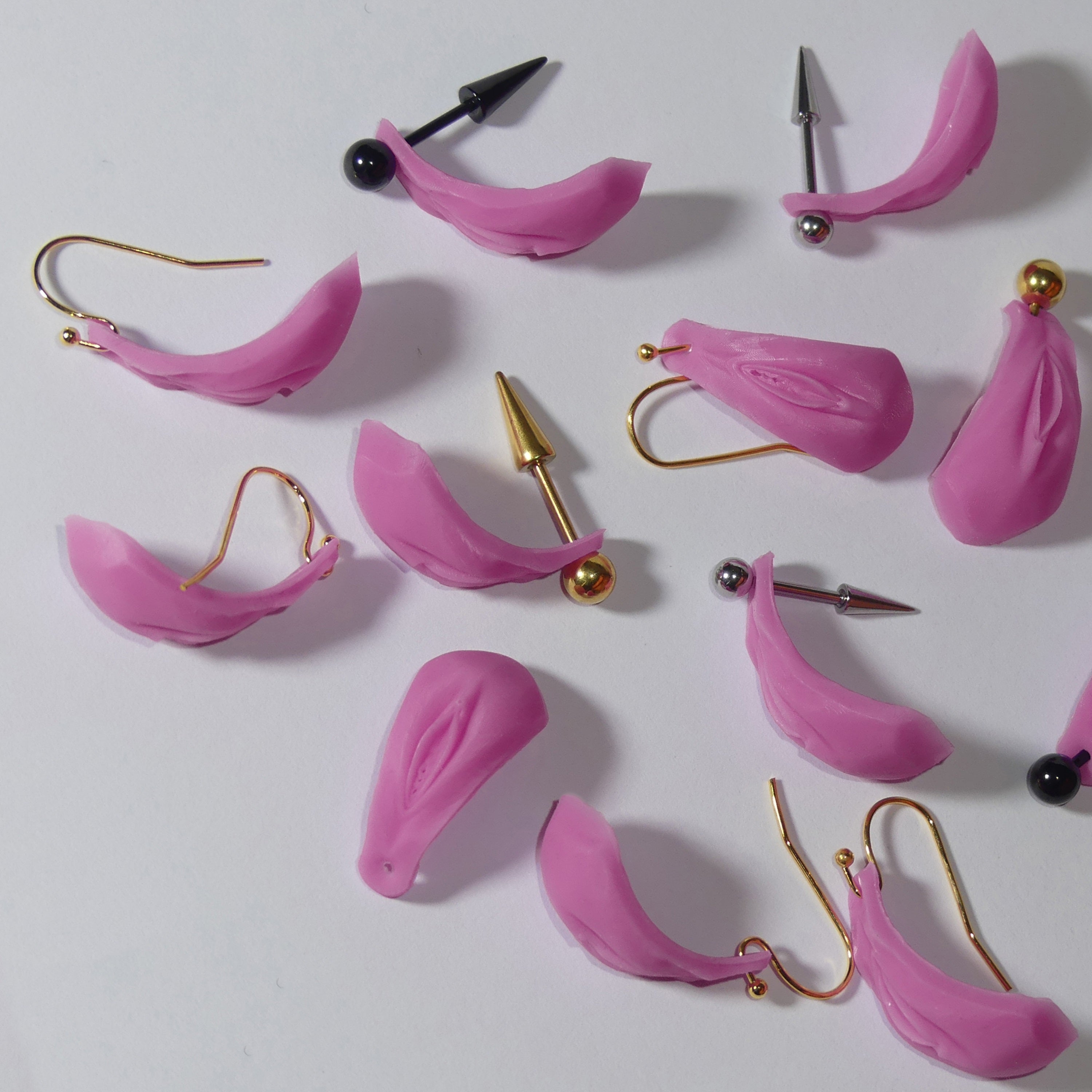 A collection of pink silicone vulva earrings with various metal ear hooks, including gold, silver, and black options, arranged on a plain white surface.