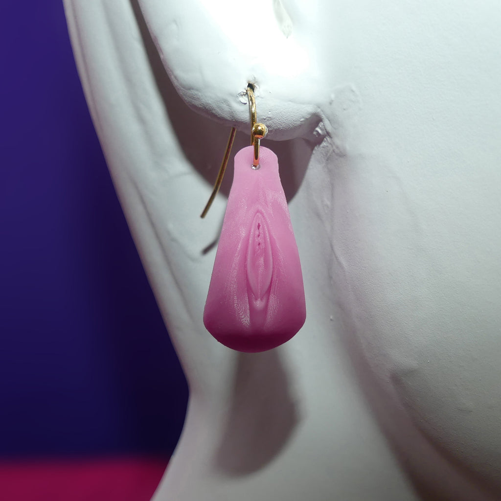A pink vulva with clitoris dangle earring with a gold fishhook wire, displayed on a white female mannequin bust against a purple and pink background.