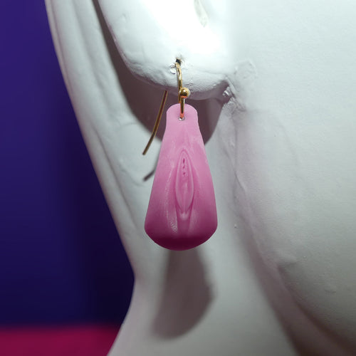 A pink vulva with clitoris dangle earring with a gold fishhook wire, displayed on a white female mannequin bust against a purple and pink background.