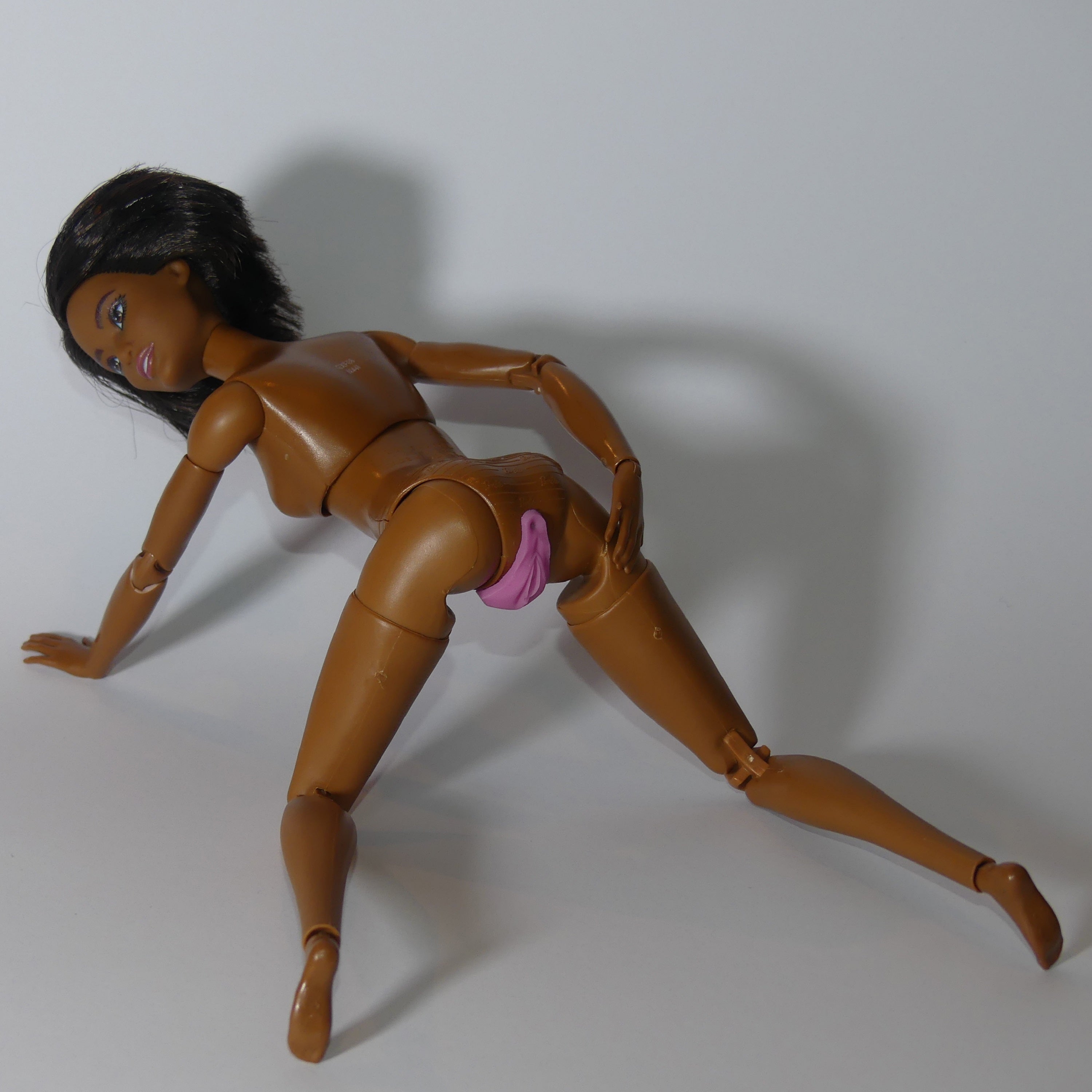 A brown articulated doll posed on a light gray background, wearing a small pink silicone vulva vagina anus under the hips.