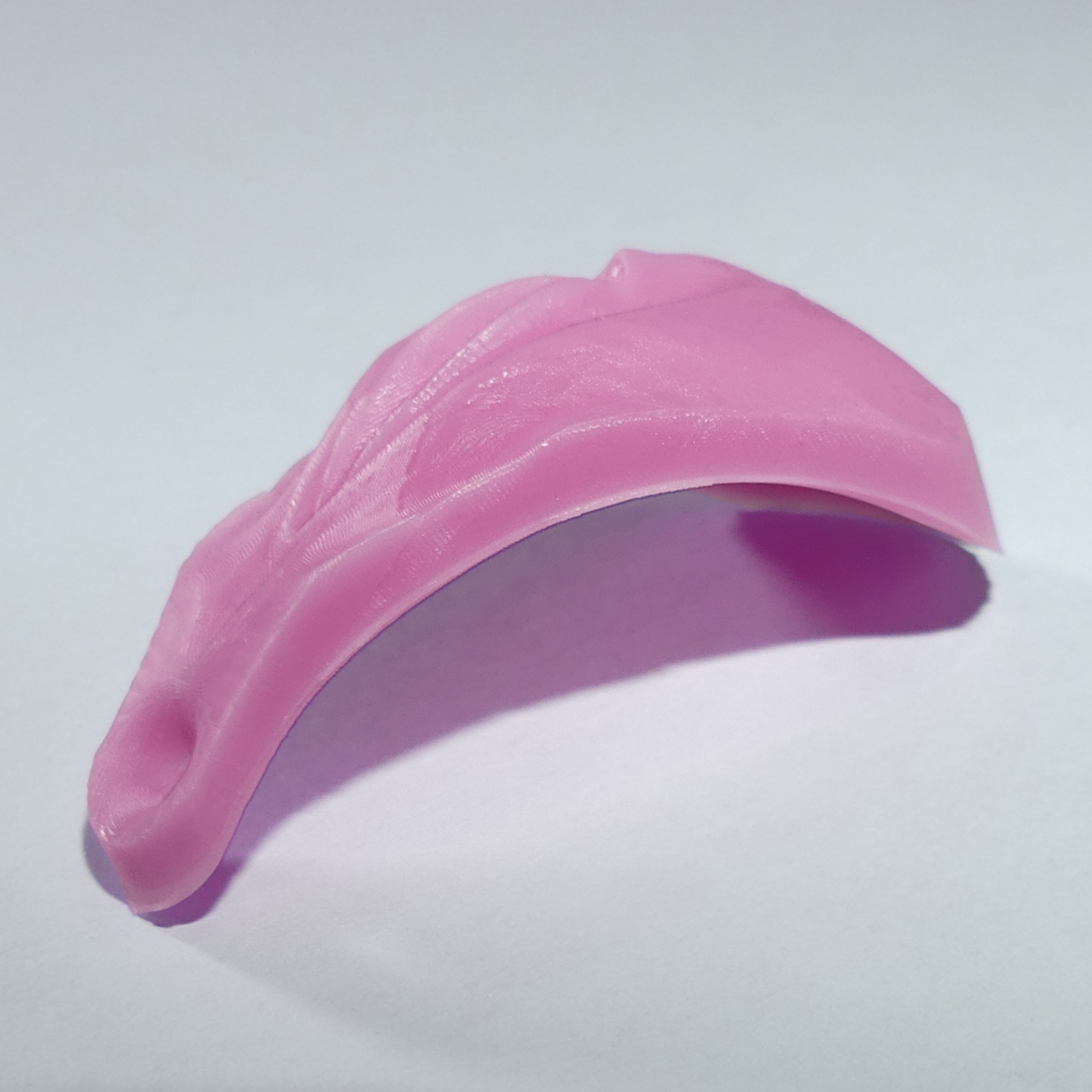 A pink, curved silicone accessory with a textured surface and a small anus at one end, shown on a plain light gray background.